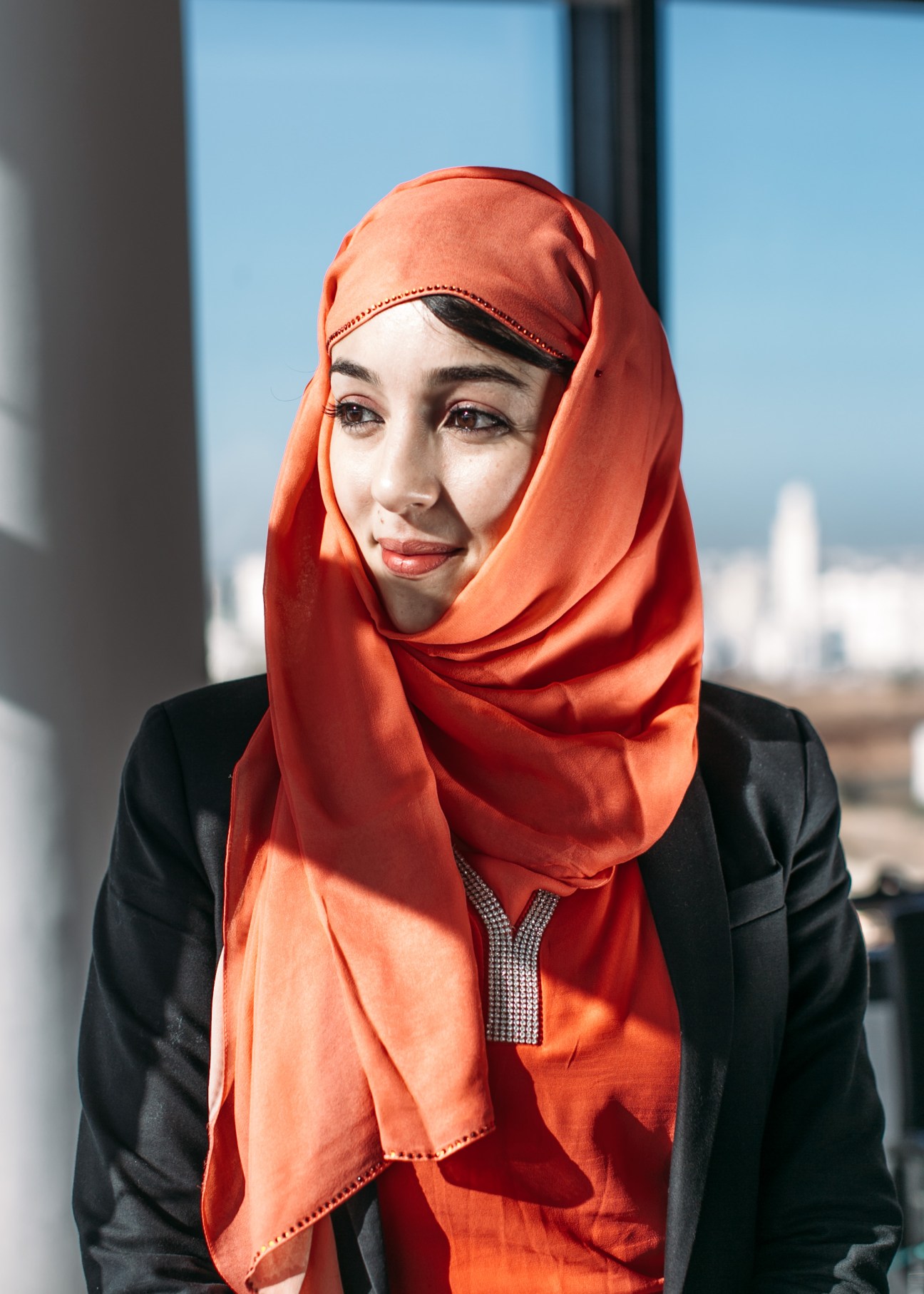 Financial-risk analyst Siham Omrana wearing a bright red hijab and black blazer against a city skyline backdrop.