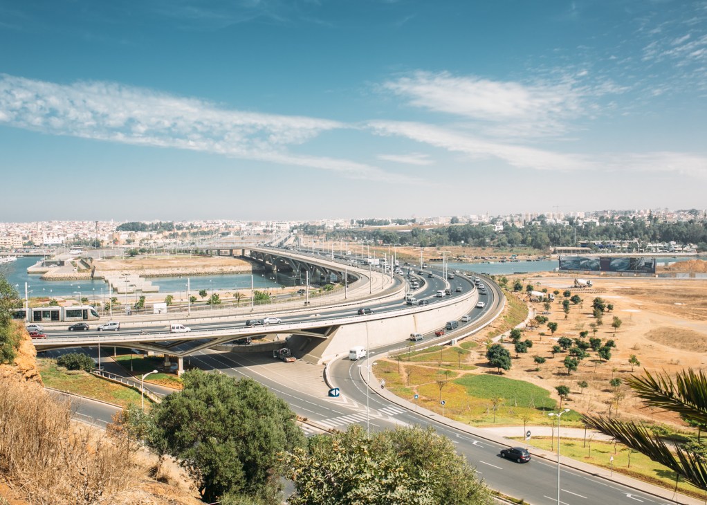 Modern highway interchange with curved ramps spanning over waterways near Rabat, Morocco, with city skyline and green areas visible.