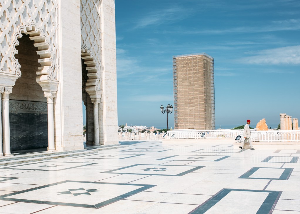Ornate white Islamic architecture with geometric patterns and carved corbels overlooks a modern high-rise building and Rabat cityscape.