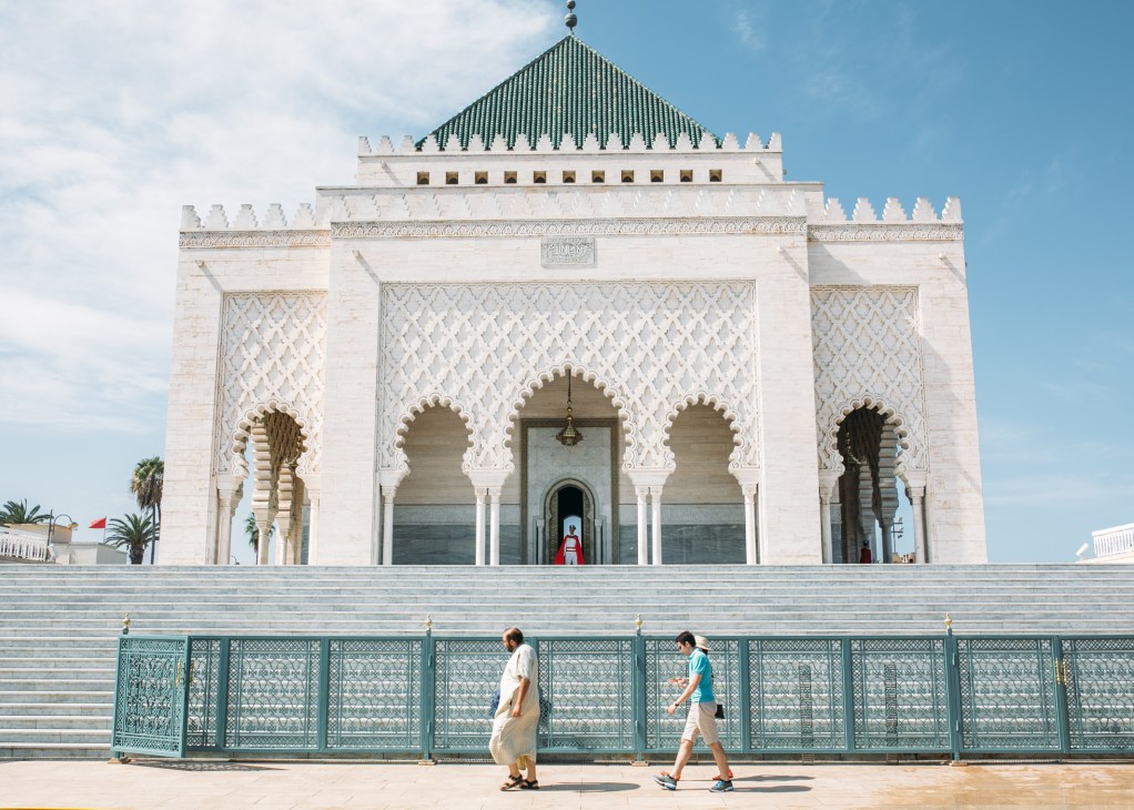 White marble mausoleum with Islamic architecture, decorative arches, and a green pyramidal roof, with visitors walking by a turquoise fence.