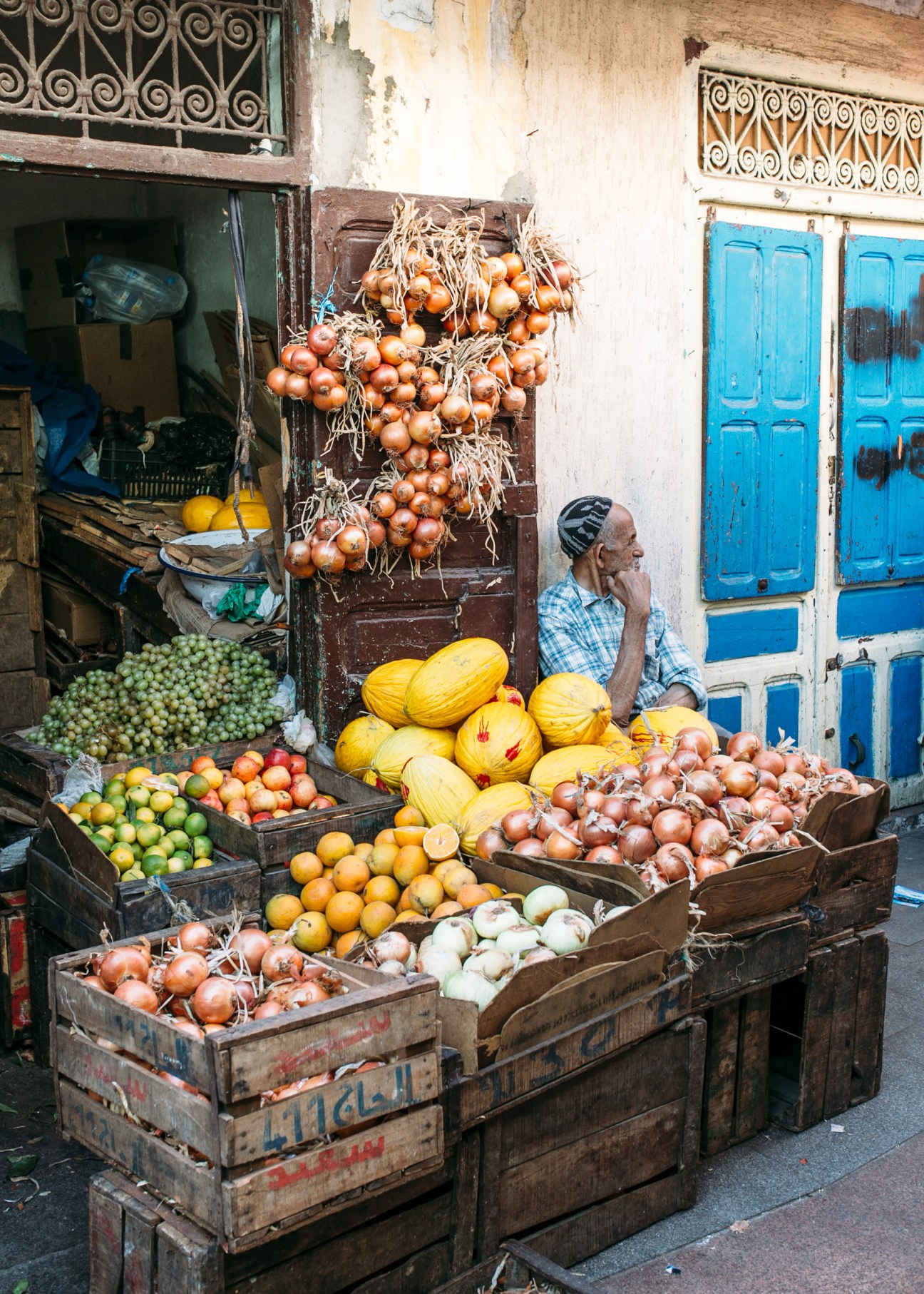 Street vendor selling fresh vegetables and onions displayed in wooden crates outside a storefront with blue doors in Rabat.