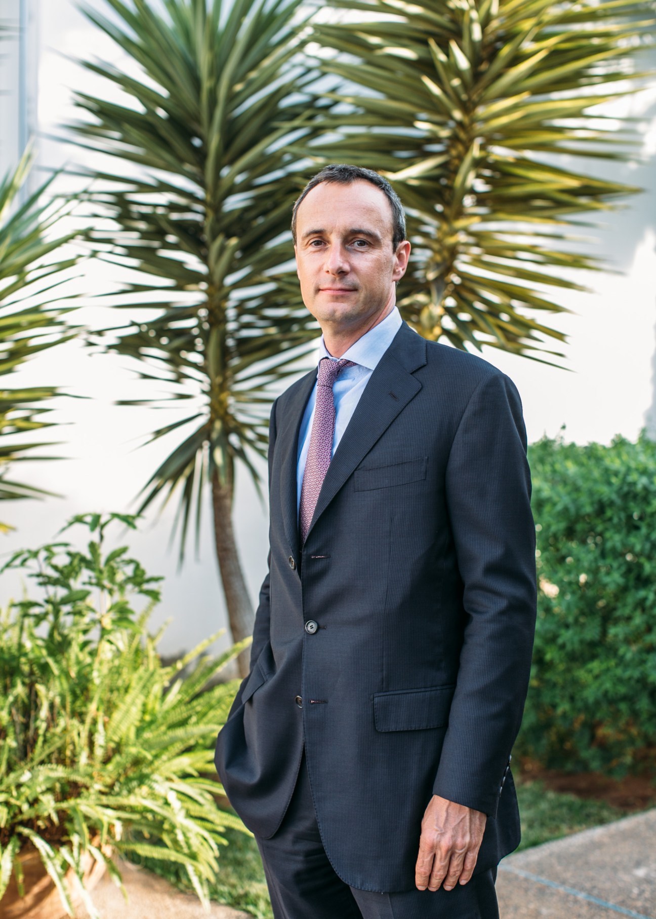 Man in dark suit and pink tie standing outdoors with palm trees in background.