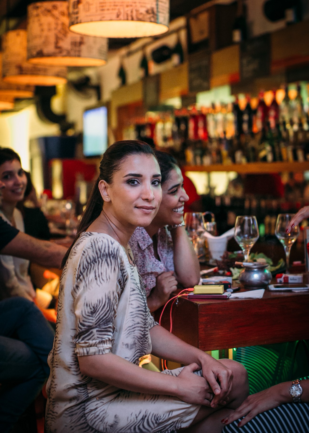 Woman in patterned dress sits at bar counter with friend, warm lighting and bottles visible in background.