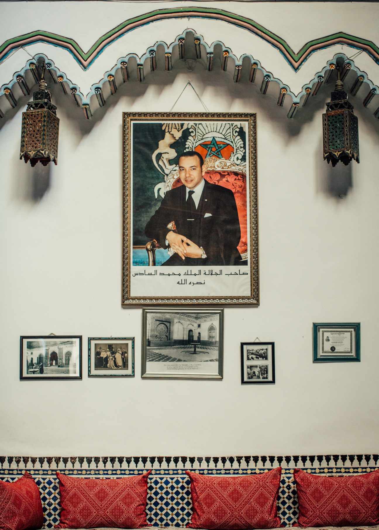 Formal portrait of King Mohammed VI in a dark suit hanging on a white wall with decorative lanterns and smaller framed photographs below.