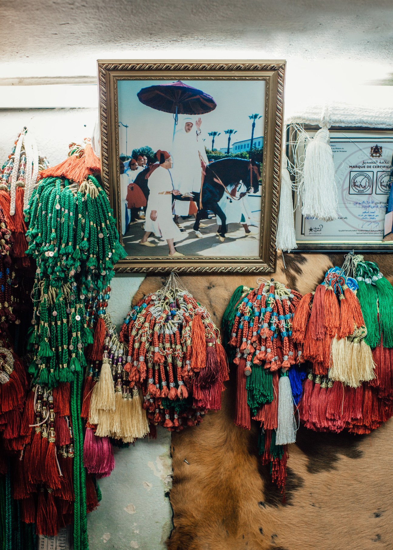 Framed photograph of King Mohammed VI with people under an umbrella displayed in a traditional saddle shop with colorful tassels.