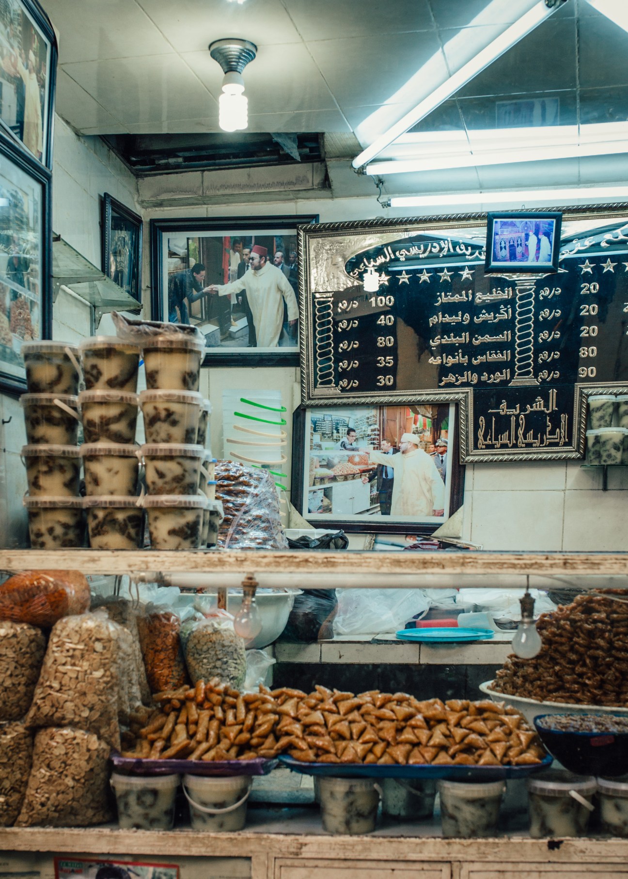 A snack bar stall in Fes souk displaying pastries and drinks with Mohammed VI portraits on the wall.