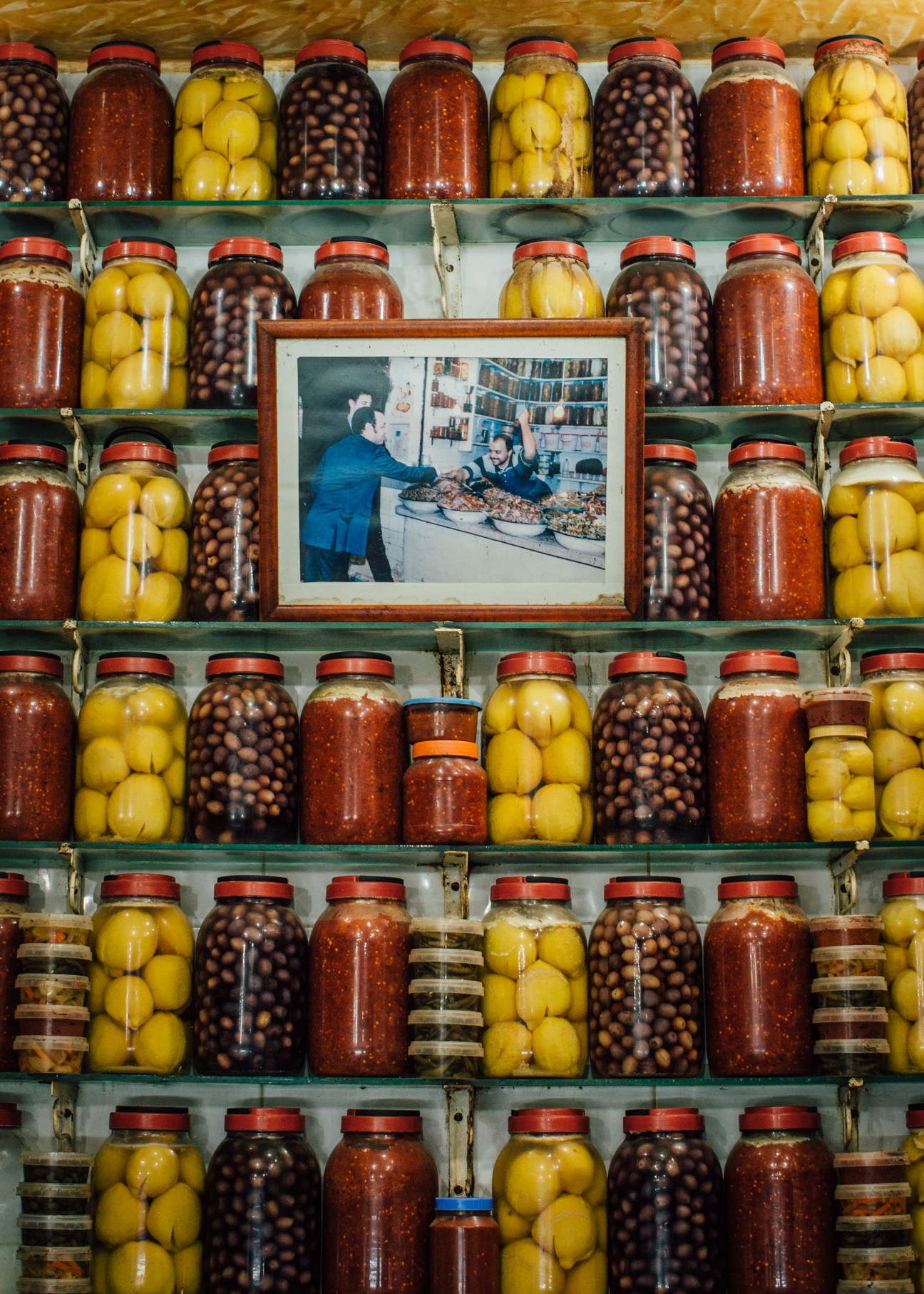 King Mohammed VI portrait in wooden frame surrounded by shelves of pickled lemons and olives in jars.