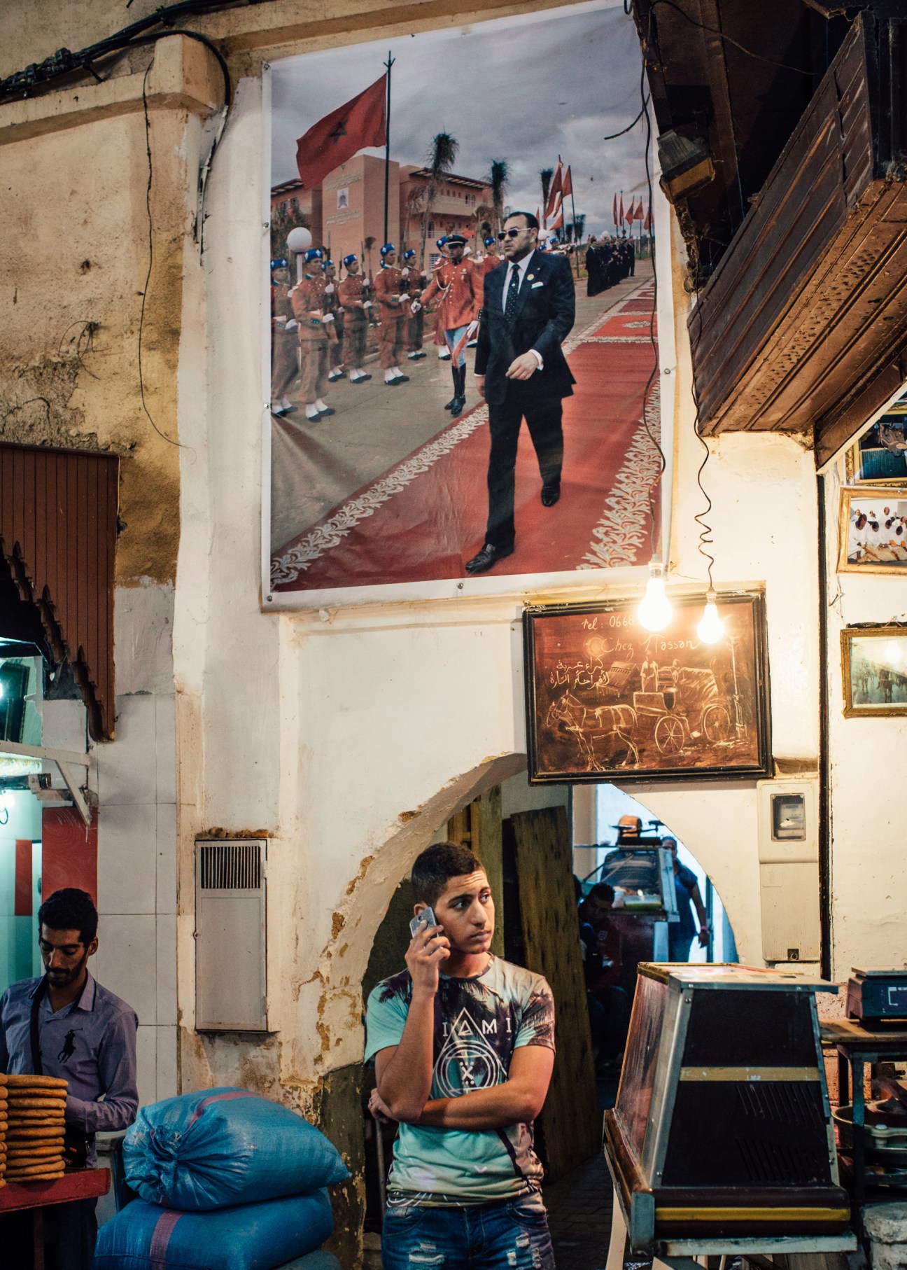 Man on phone in Fes souk beneath official portrait of King Mohammed VI.