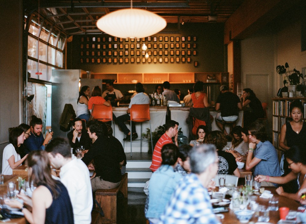 Diners seated at communal tables enjoying meals at Stray Dogs restaurant with orange chairs and industrial ceiling.