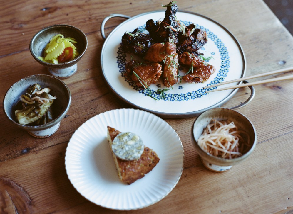 Korean fried chicken with corn bread and pickles served on white plates with small bowls of side dishes on wooden table.