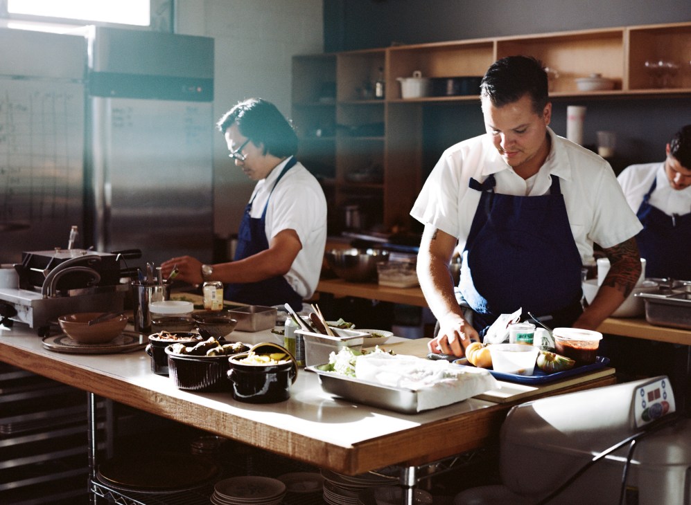Two chefs in white shirts and dark aprons prepare food at a kitchen counter.