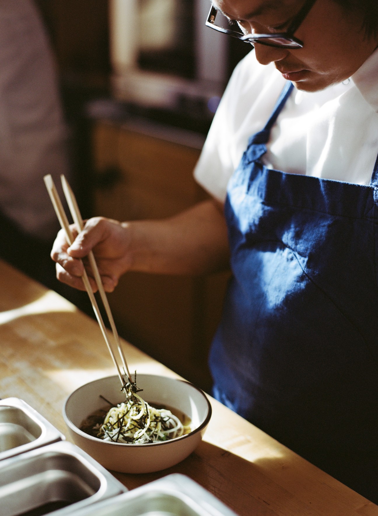 Chef Peter Cho applies final touches to soba noodles with chopsticks in a white bowl.