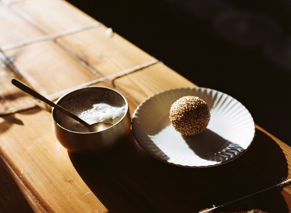 Sesame ball with shaved Vietnamese iced coffee on a white plate and dark bowl on wooden surface.