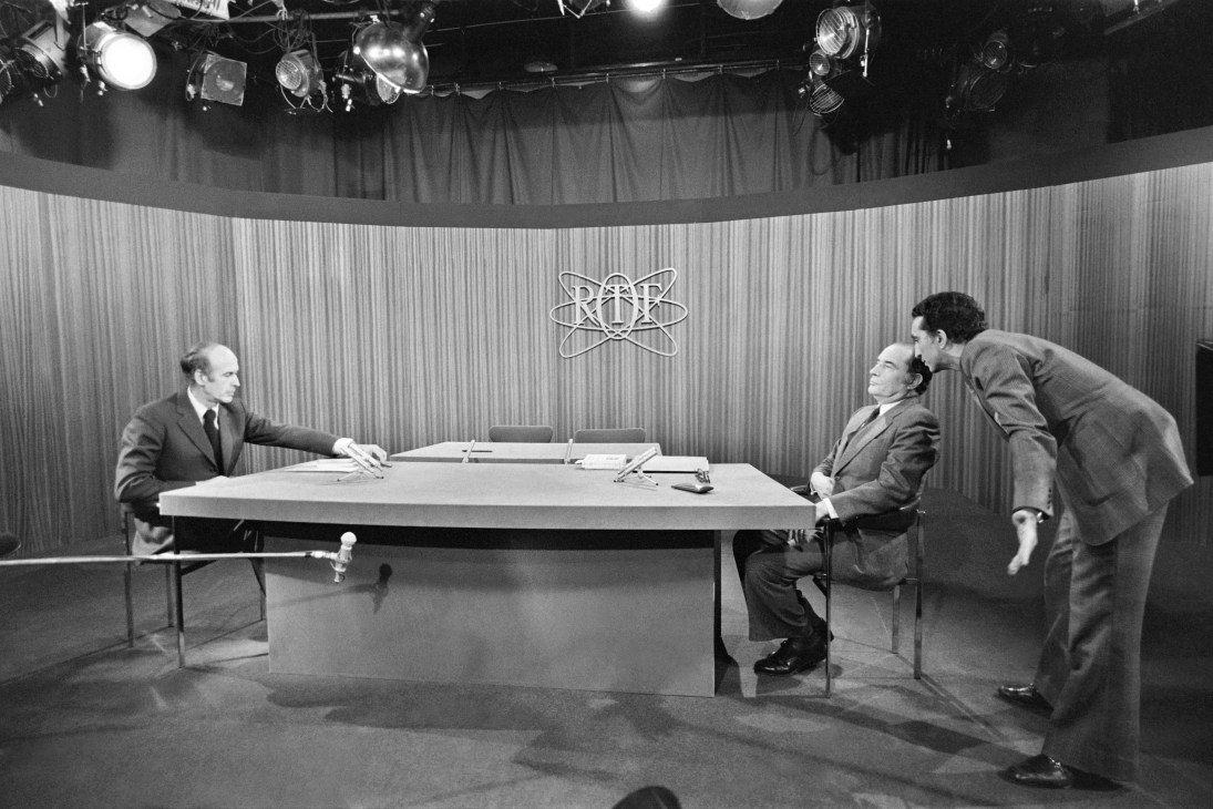 Black and white photograph of two men at a table on a television set with RTF logo visible on the backdrop.