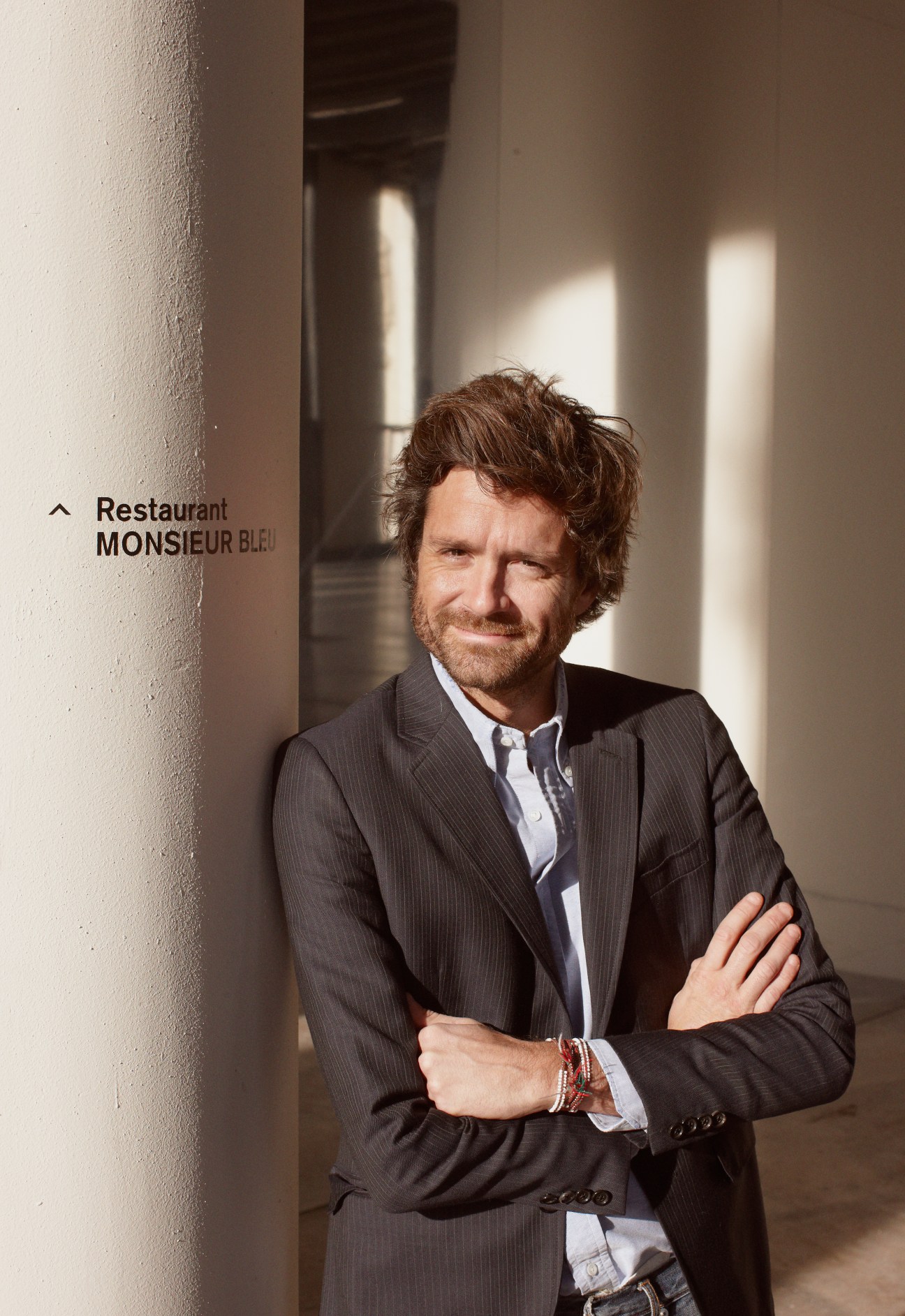 Architect Joseph Dirand stands in a minimalist corridor next to Restaurant Monsieur Bleu signage.