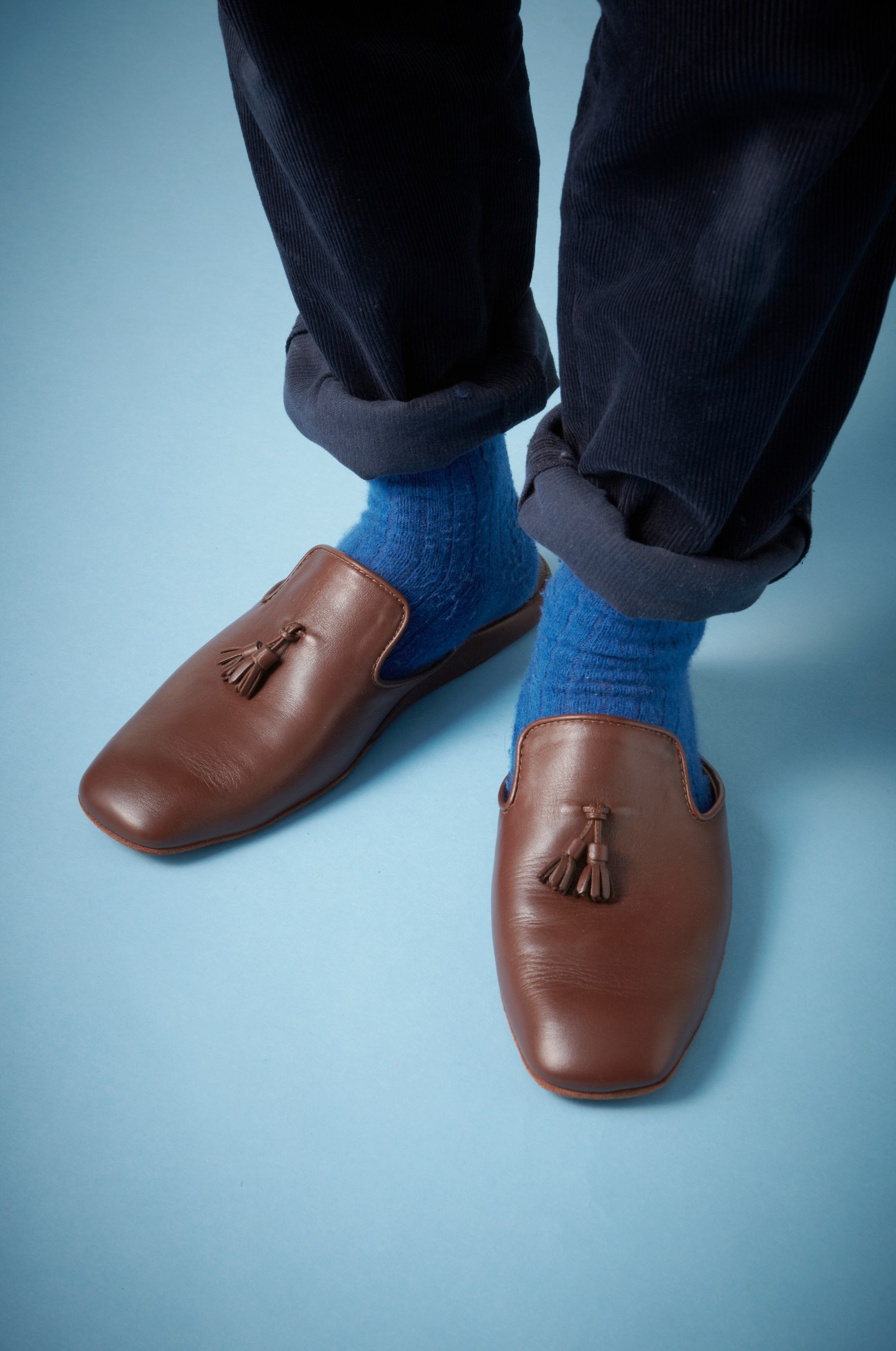 Brown leather slip-on slippers with tassels worn with blue socks and rolled navy trousers against a light blue background.