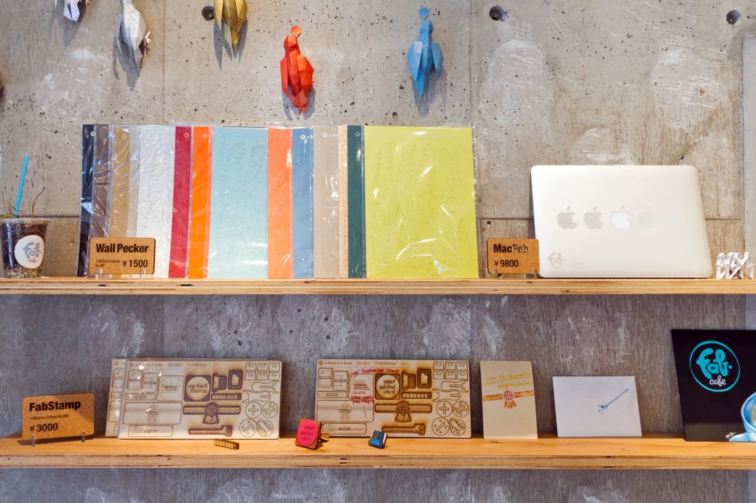 Interior shelving at FabCafe Tokyo displaying colorful notebooks, laptop, and laser-cut wooden design samples.
