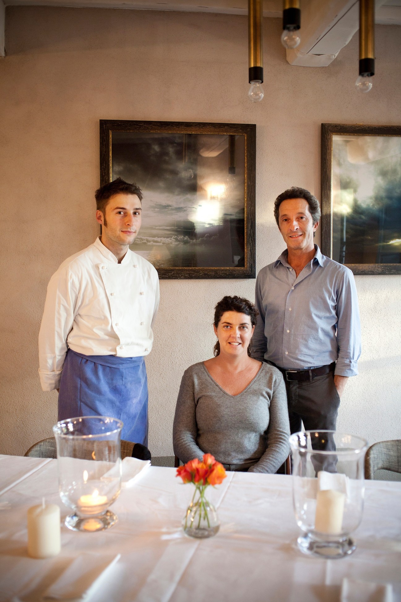 Three restaurant staff members posing in an elegant dining room with framed artwork on walls.