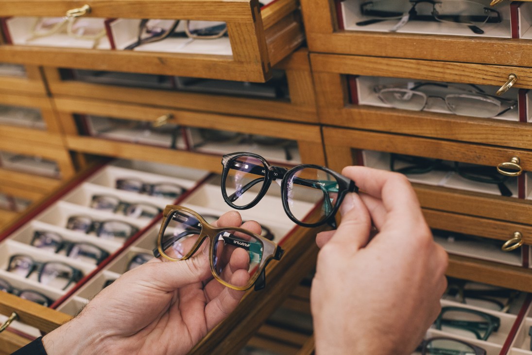 Hands holding two pairs of custom horn-rimmed eyeglasses inside a wooden display case at Hartmann optician.