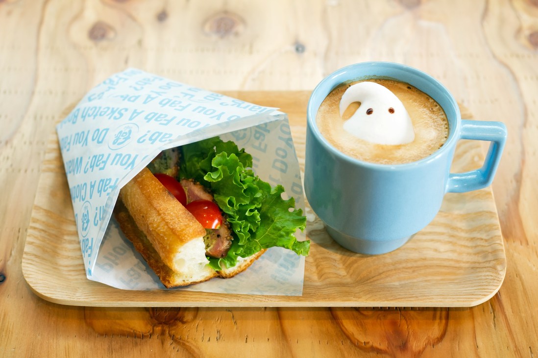 A sandwich wrapped in blue paper and a blue mug with latte art featuring a ghost design on a wooden board.