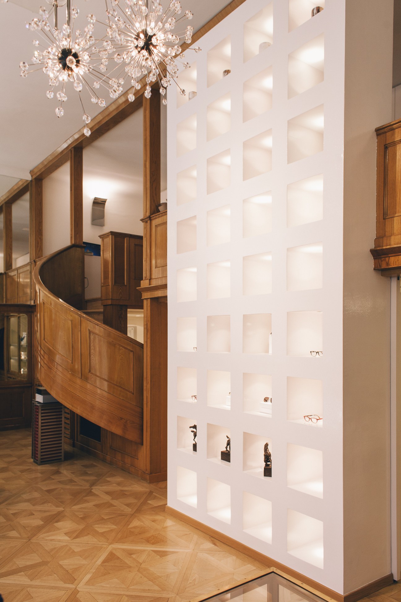 Interior of Hartmann eyewear shop showing wooden spiral staircase, crystal chandelier, and white wall display niches.