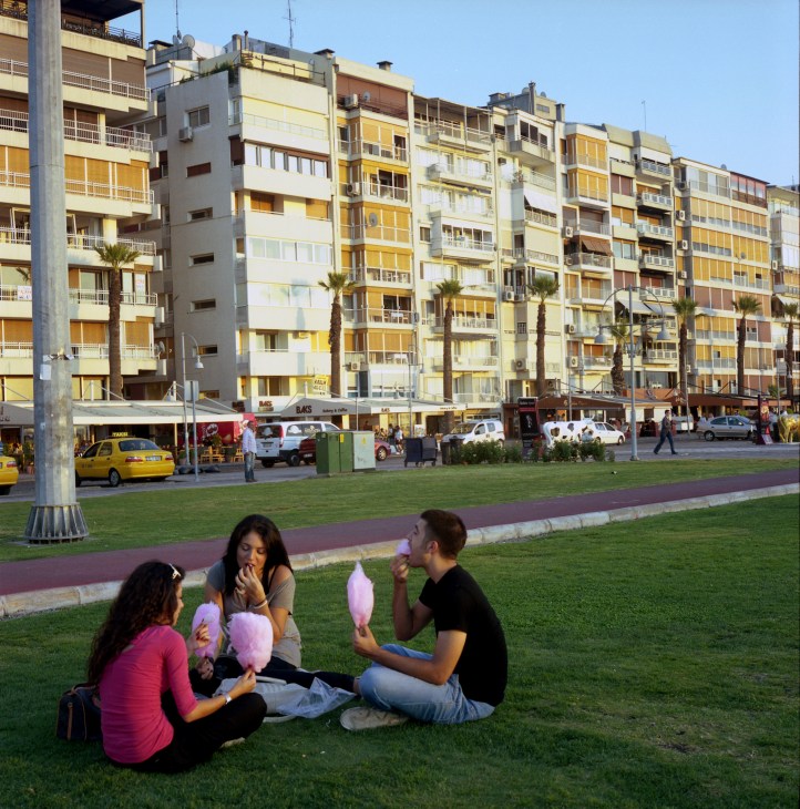 Young people eating candyfloss on a grassy waterfront promenade with modern apartment buildings behind them in Izmir.