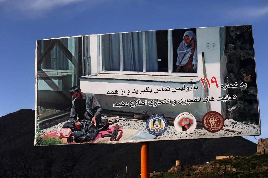 Afghan government anti-terrorism billboard with Dari and Pashtu text warning against explosive devices and insurgent activities.
