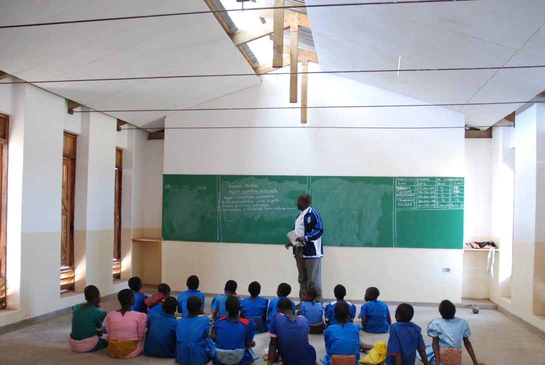 A teacher stands at a chalkboard instructing students seated on the floor in a bright classroom with skylights.