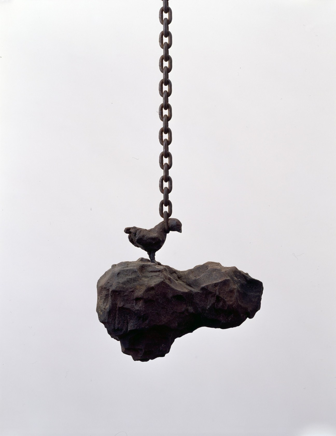 Small bird sculpture perched on a dark rock suspended by a metal chain against white background.
