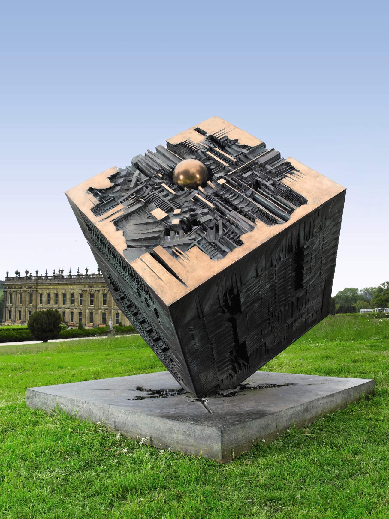 Large abstract bronze and steel sculpture displayed on a concrete base in manicured gardens with a grand estate house in the background.