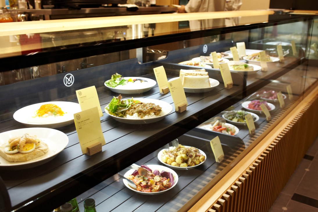 Display case of Monocle Café menu items including pork cutlet sandwiches, Swedish skagen, and Reuben sandwiches with price cards.