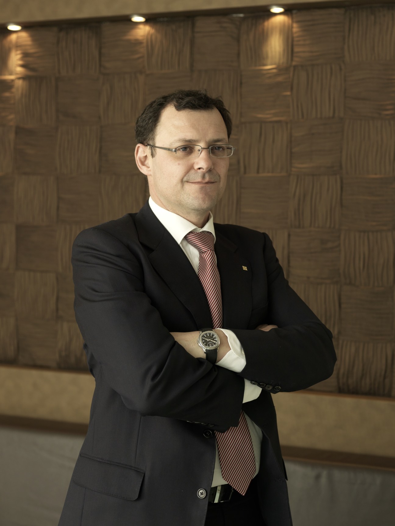 Thierry Stern, president of Patek Philippe, wearing a dark suit and striped tie with a watch visible on his wrist, standing with arms crossed against a textured wall.