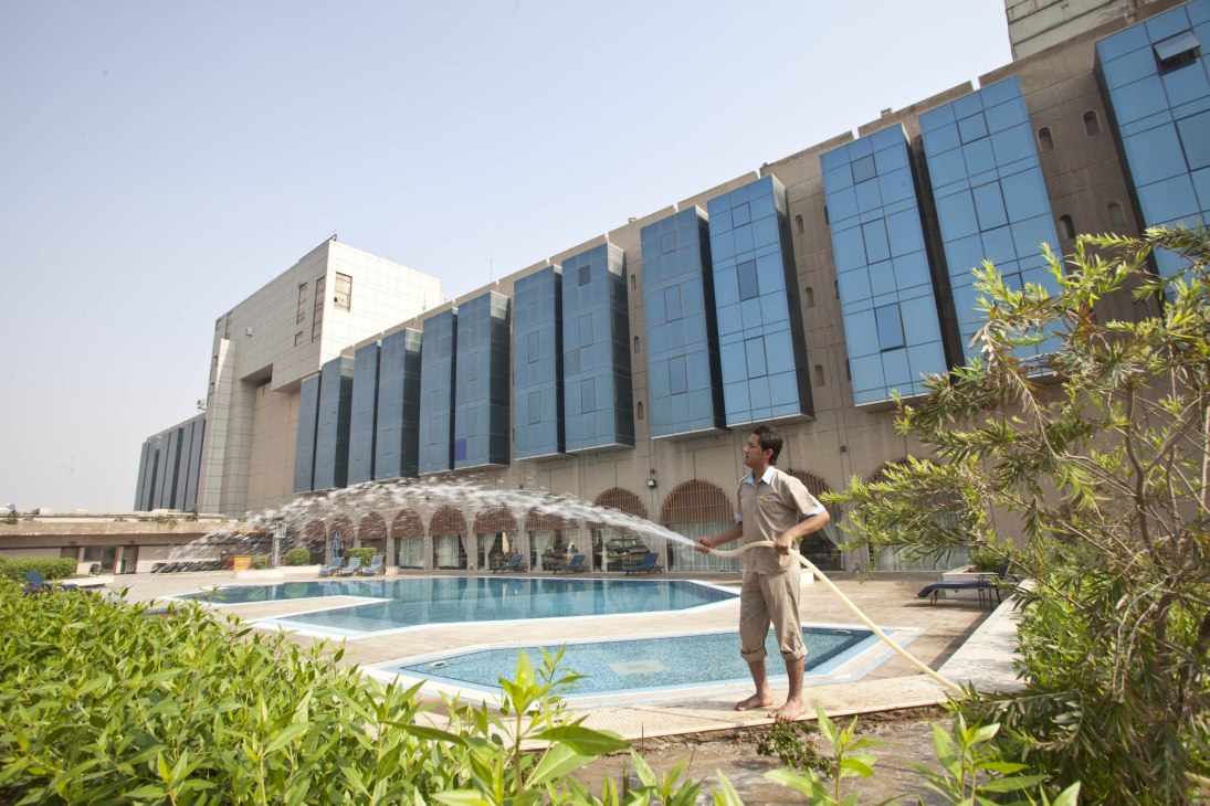A gardener waters plants in front of the Basra International Hotel's modern glass and concrete building with swimming pools.