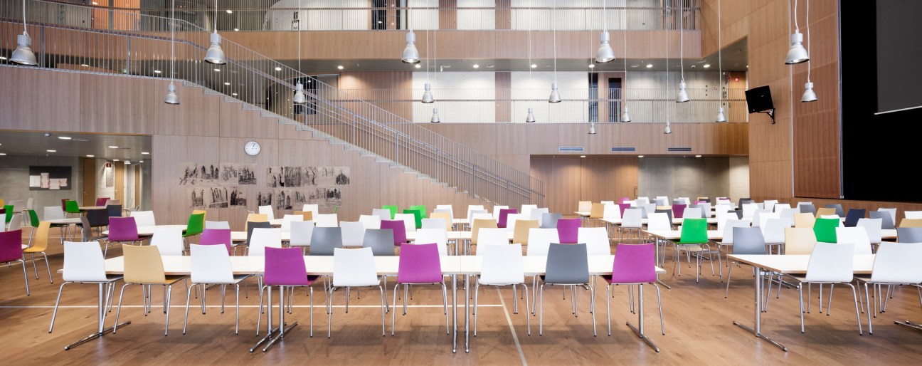 Modern school canteen with oak wood interior, colorful chairs, and pendant lighting under high ceiling with mezzanine level.
