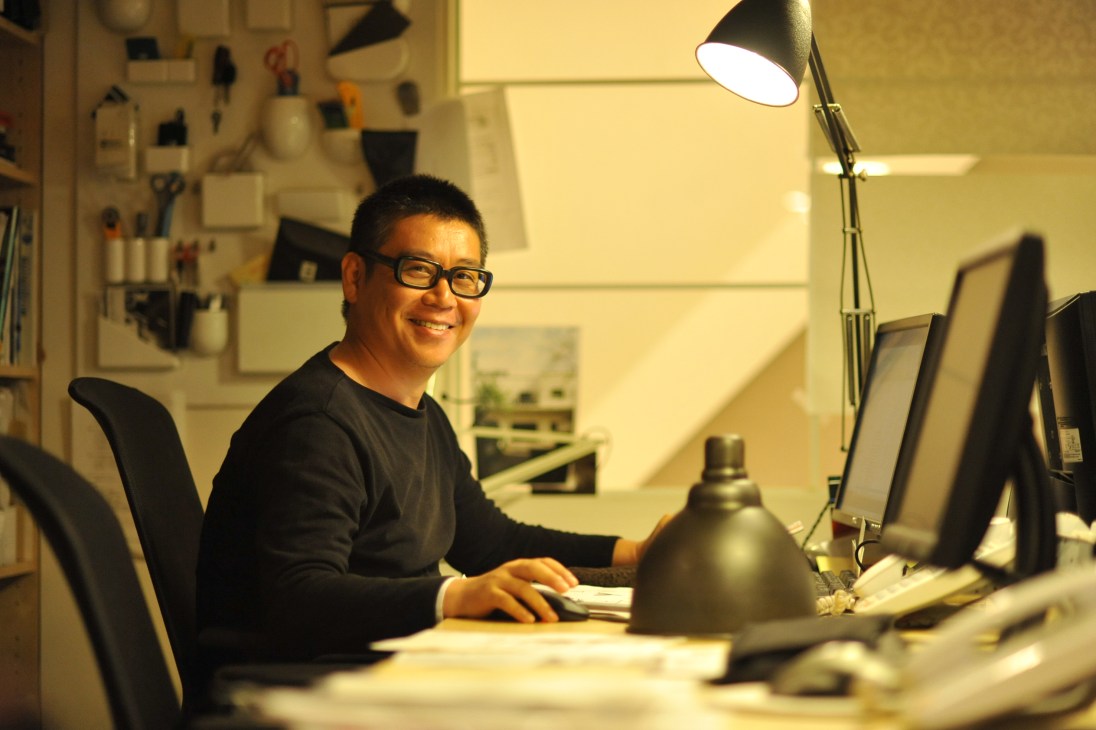 Lighting designer Kenichi Kandatsu smiles at his desk in his Osaka studio, surrounded by design sketches and a brass pendant light.