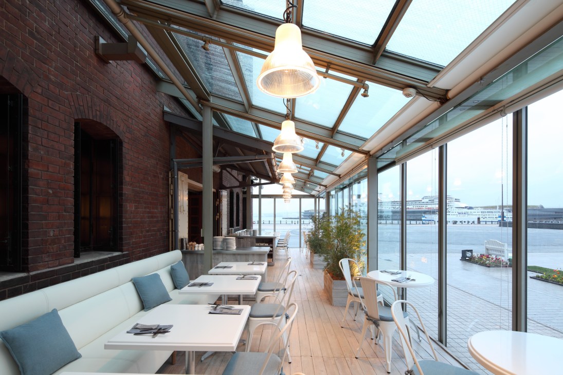 Bright modern restaurant interior with glass ceiling, pendant lights, white seating and waterfront views.