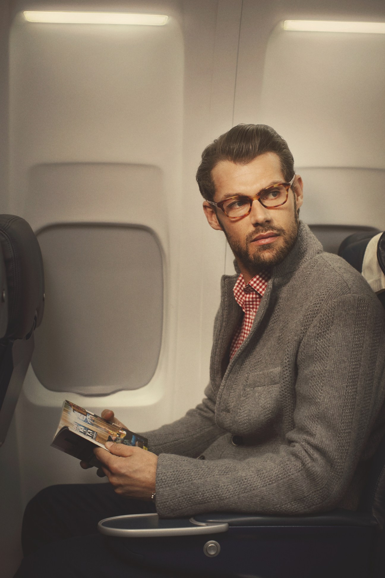Man wearing Prada glasses and Barena jacket on an airplane, holding a magazine