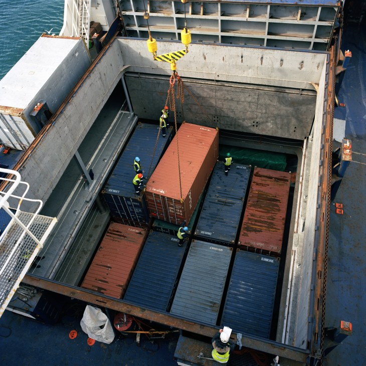Workers in high-visibility vests load shipping containers into the cargo hold of a ship using a yellow crane.