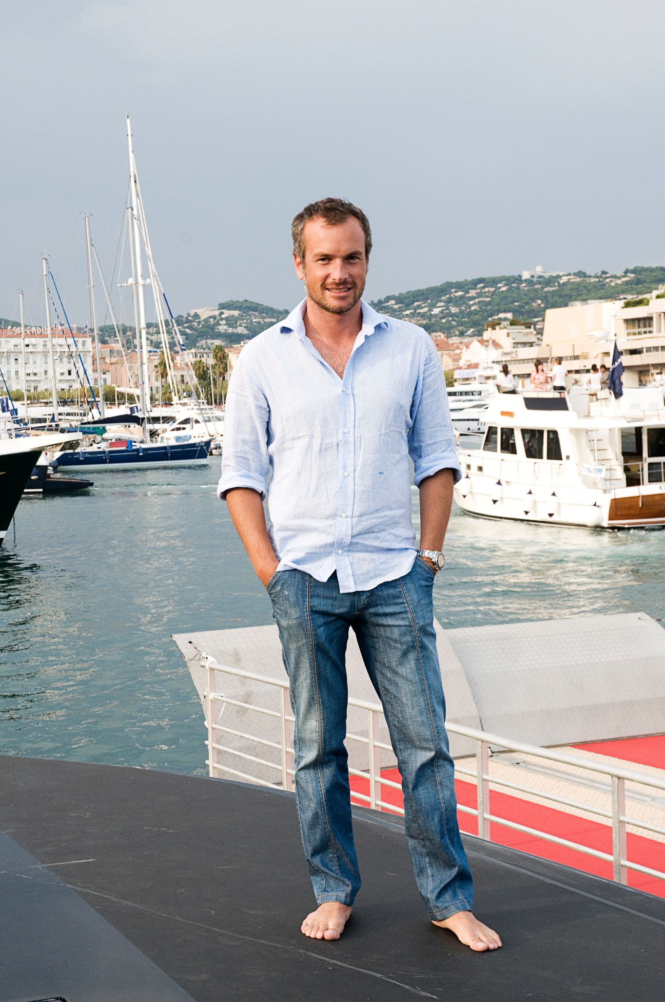 Marco Meneghini stands barefoot on a boat deck in Cannes, with sailboats and Mediterranean buildings behind him.