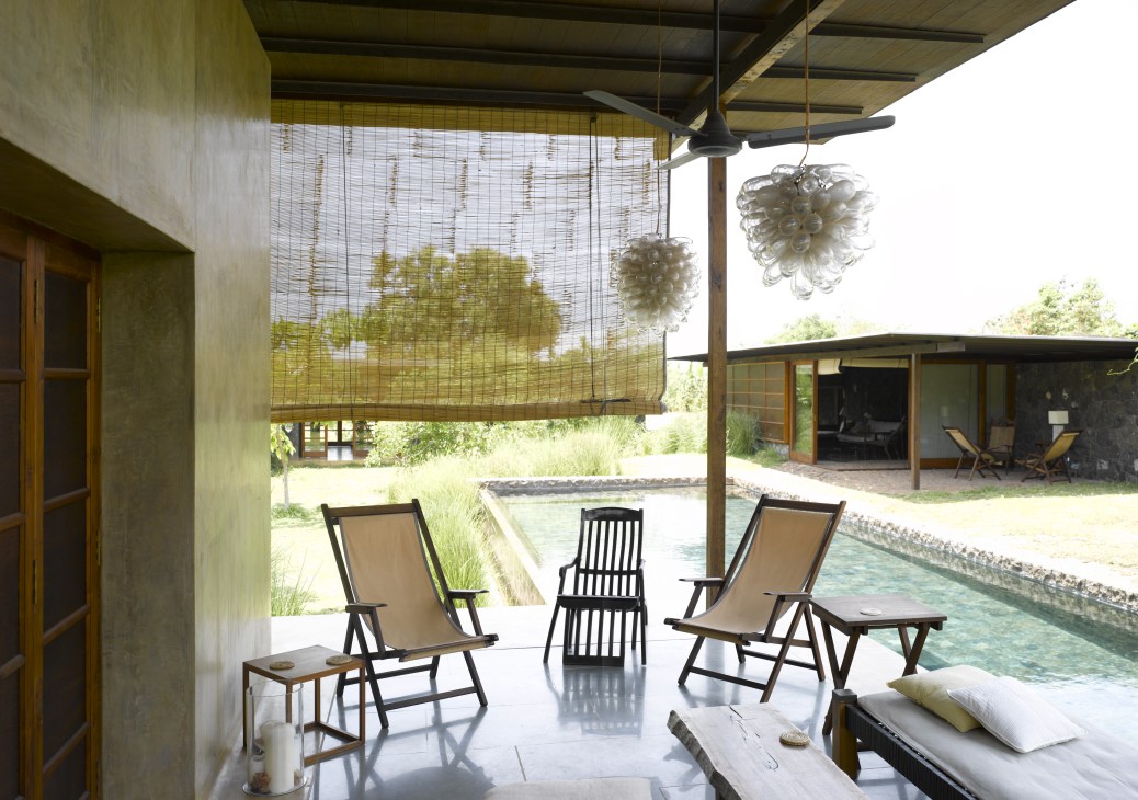 Covered terrace with lounge chairs overlooking a pool and landscape, featuring woven screens and hanging light fixtures.