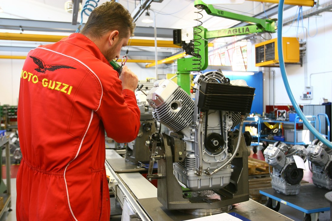 Worker in red Moto Guzzi jacket assembles motorcycle engine in factory workshop.