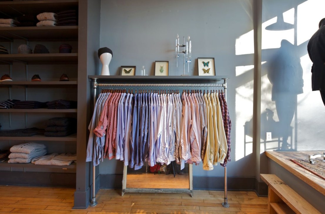 Oliver Spencer boutique interior with clothing rack displaying shirts in various colors and wooden shelving with folded items.