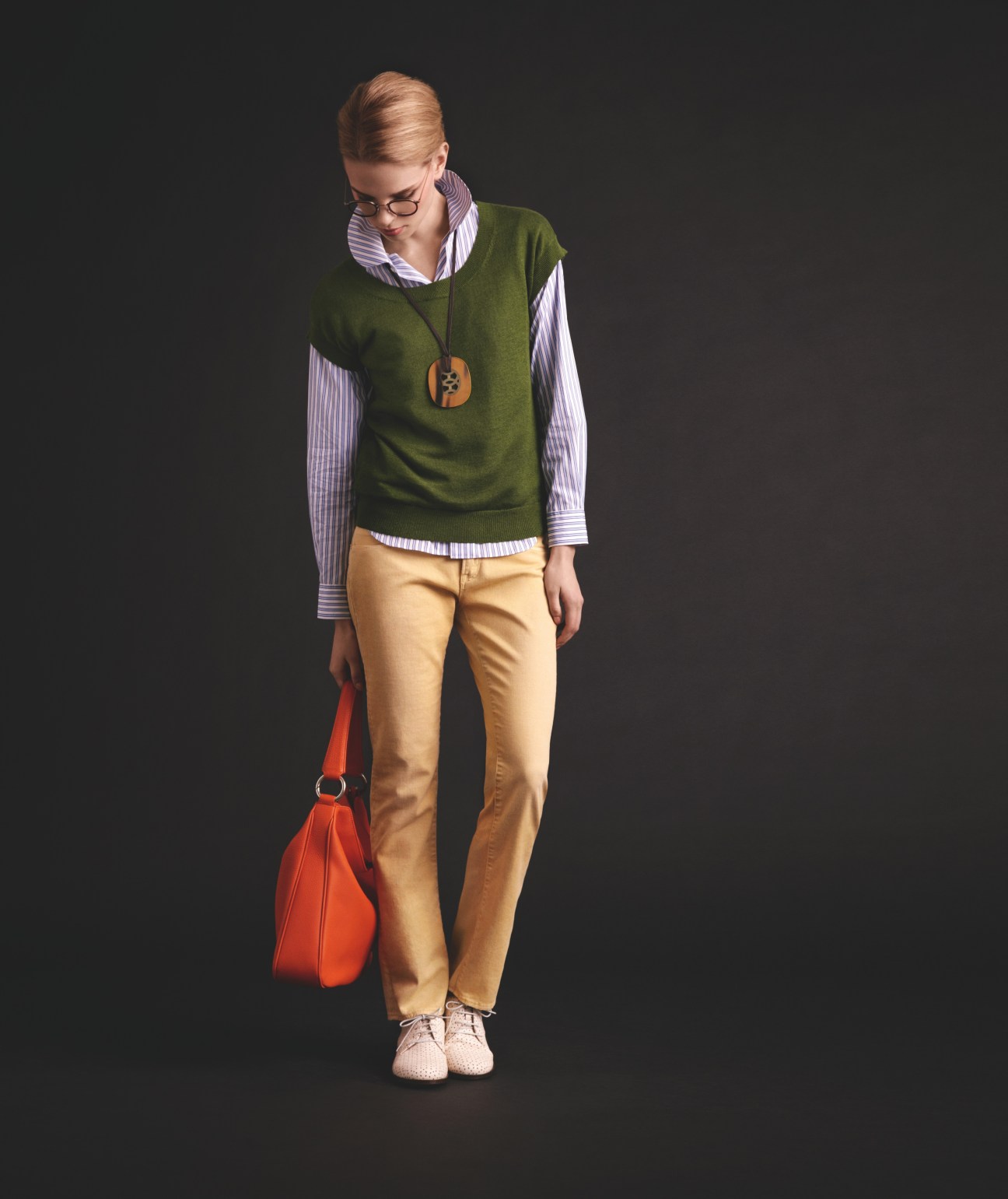 Model wearing Oliver Peoples glasses, green sleeveless top, striped shirt, Hermès necklace, beige trousers, and carrying a red bag.