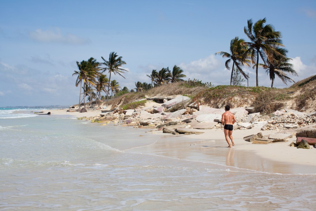 A man wades into turquoise water at a sandy beach lined with palm trees and rocky outcrops in Guanabo, Cuba.