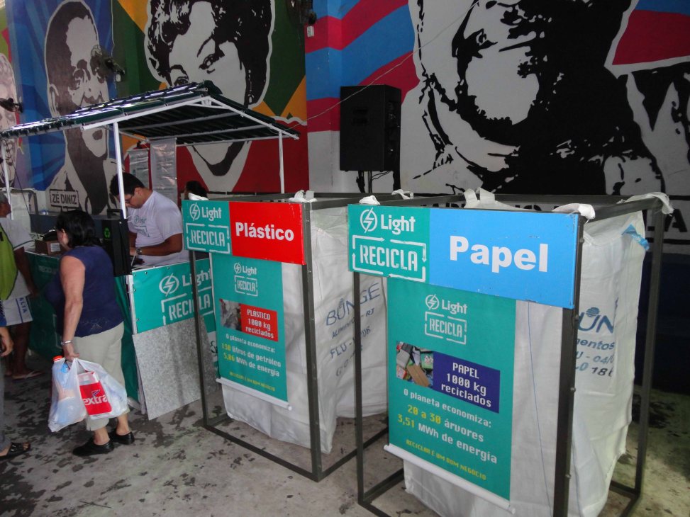 Recycling collection bins labeled "Light Recicla" for plastic and paper in a Brazilian favela street.