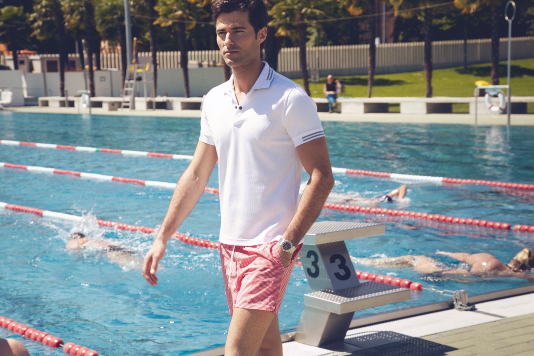 Man in white polo shirt and red patterned swim trunks standing by a swimming pool with lanes