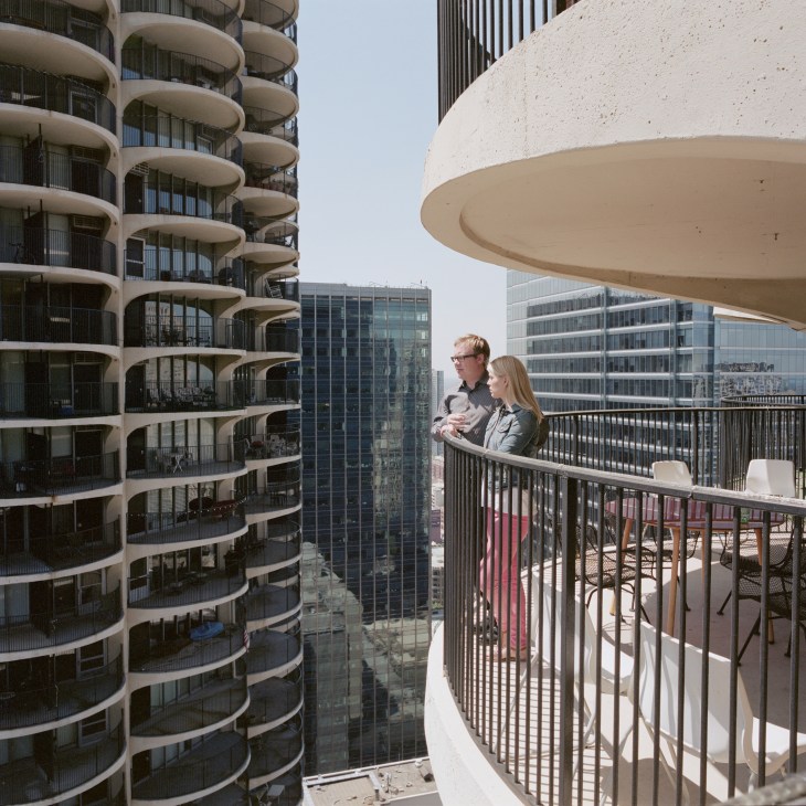 A man and woman stand on a curved concrete balcony of Marina City's East Tower, overlooking Chicago's skyline and the scalloped cylindrical tower beside them.
