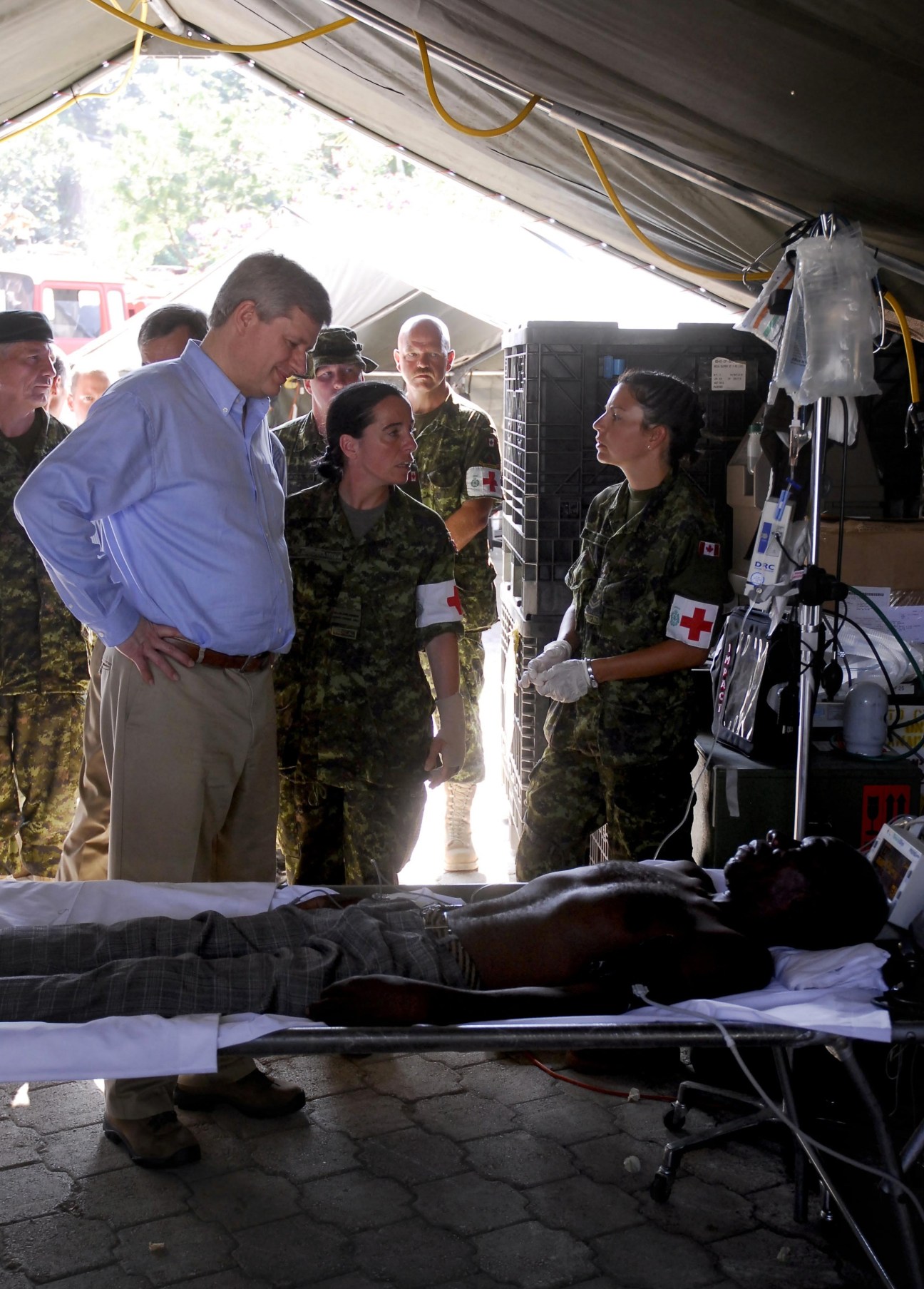 Prime Minister Harper in a blue shirt speaks with Canadian military personnel wearing camouflage uniforms inside a medical tent in Haiti.