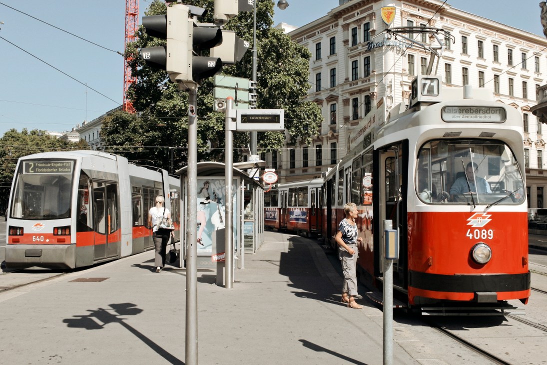 Red and white trams stop at a Vienna street station with passengers boarding and classical buildings in the background.