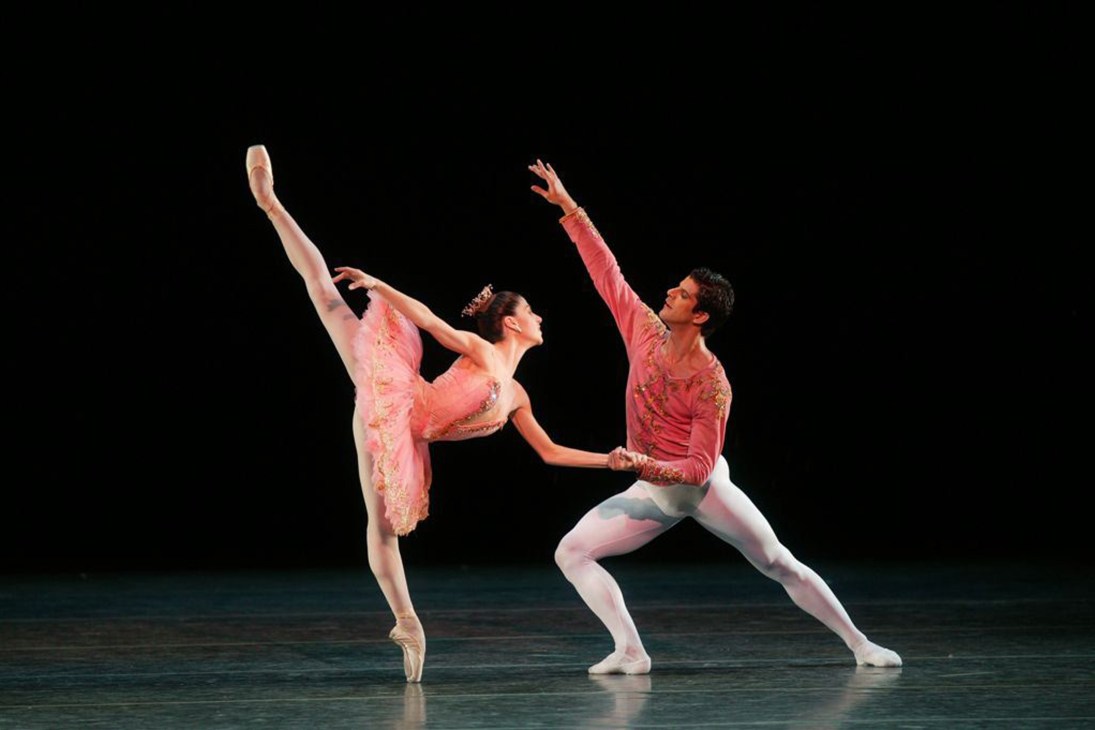Two ballet dancers perform together on stage, the female dancer in a pink tutu held in arabesque by her male partner in pink costume.